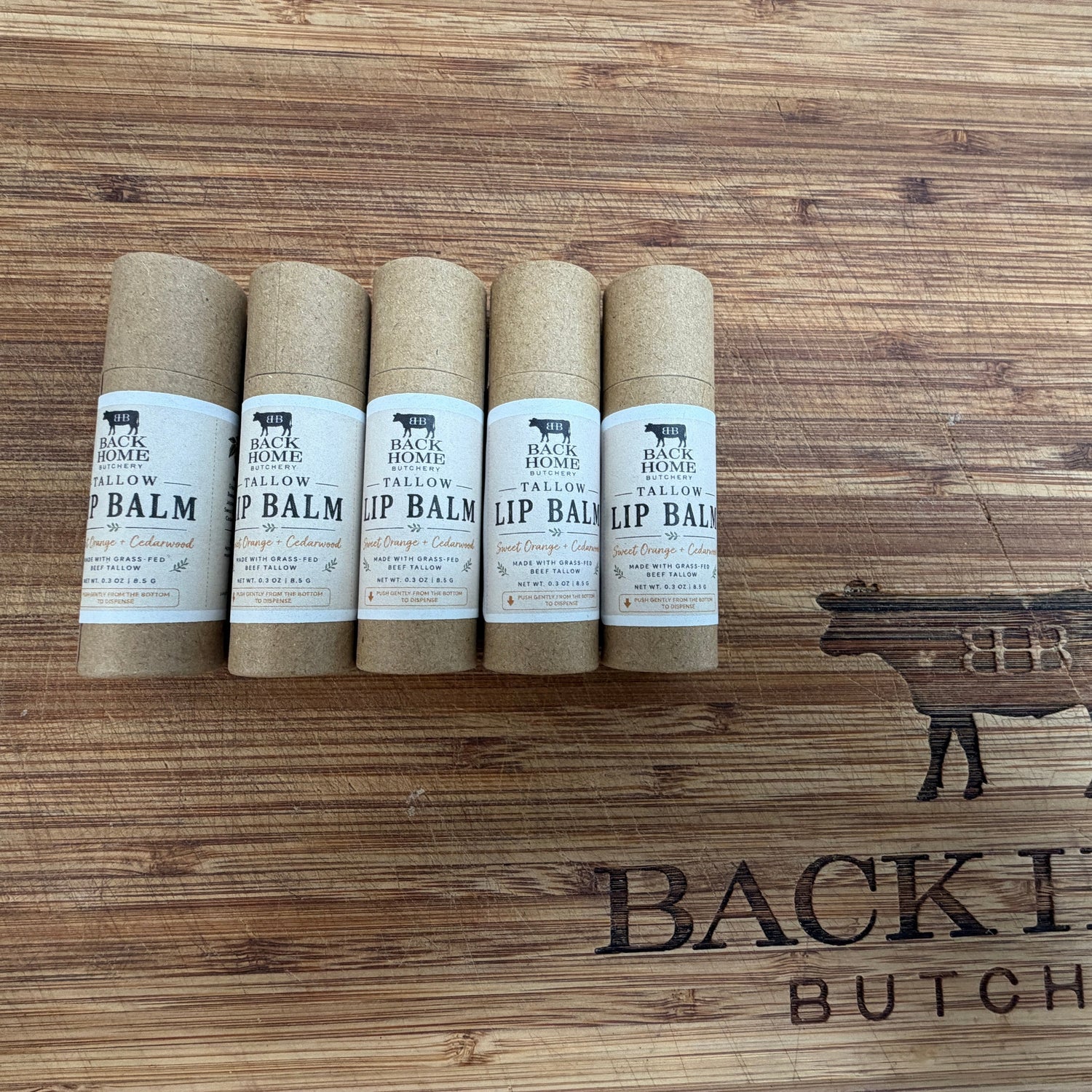 Six rolled lip balm tubes arranged vertically on a wooden surface with a visible 'BACK HOME BUTCHERY' logo engraved on the left side.